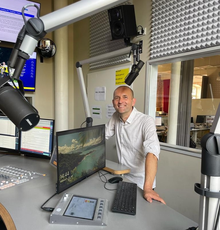 A middle-aged man (me) in a white dress shirt and jeans in a radio recording booth